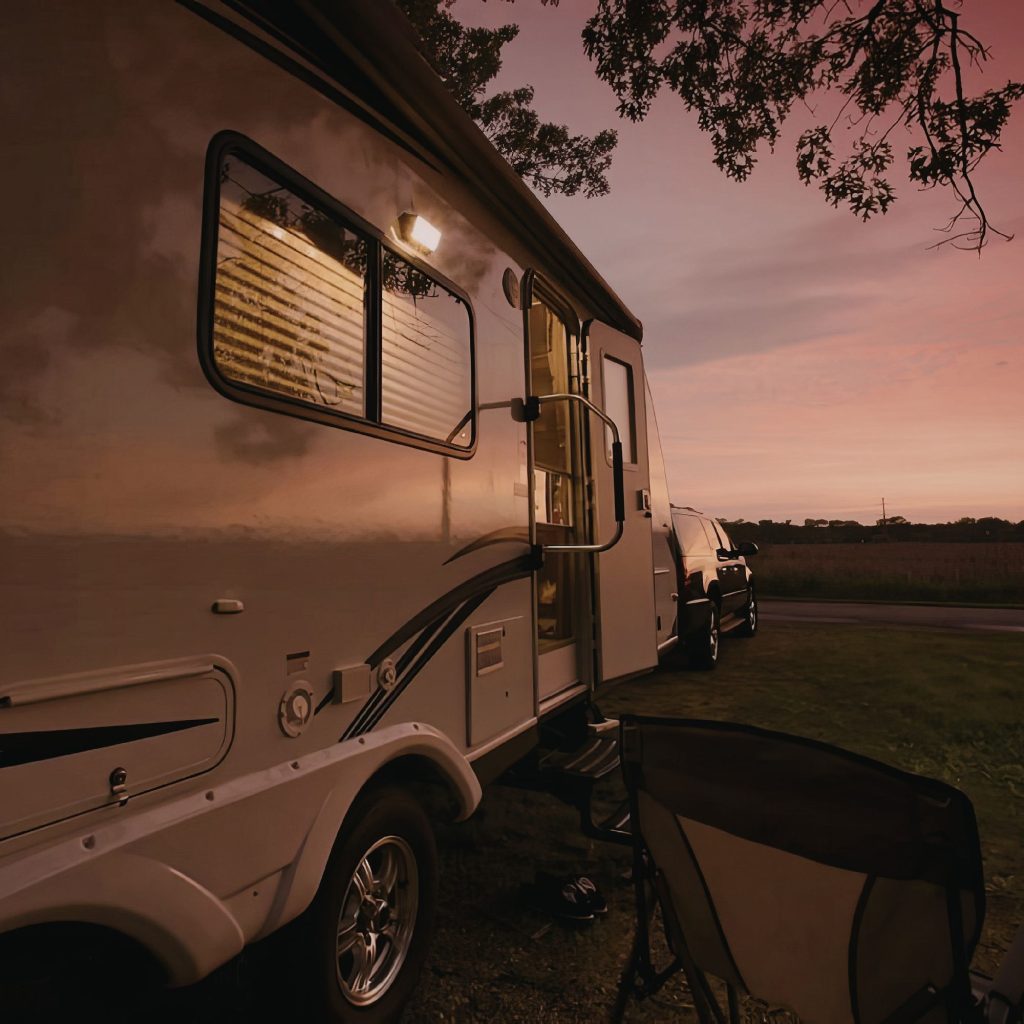 A white RV parked outdoors at sunset with pink sky, tree branches in the background and chairs on the ground nearby.