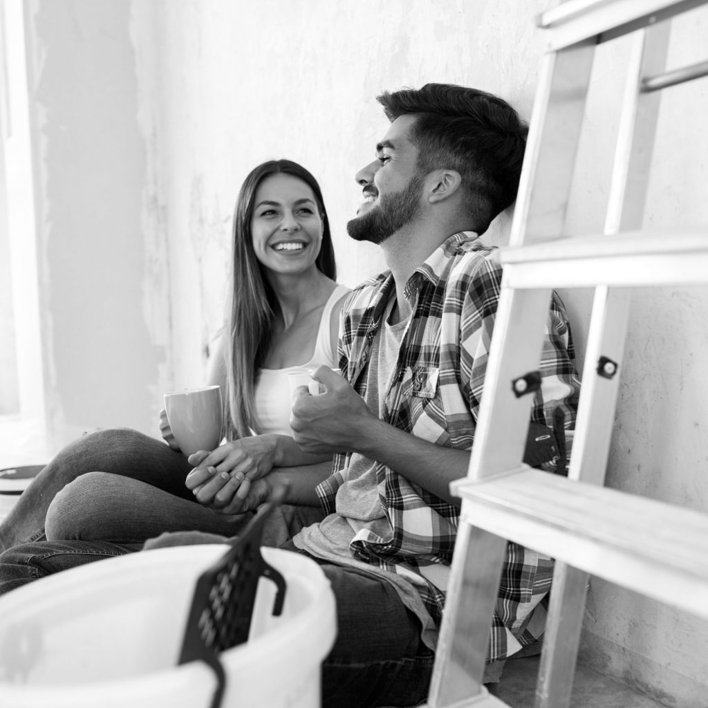 Smiling couple sitting on a bench beside a ladder, each holding a mug and chatting outdoors (black and white).