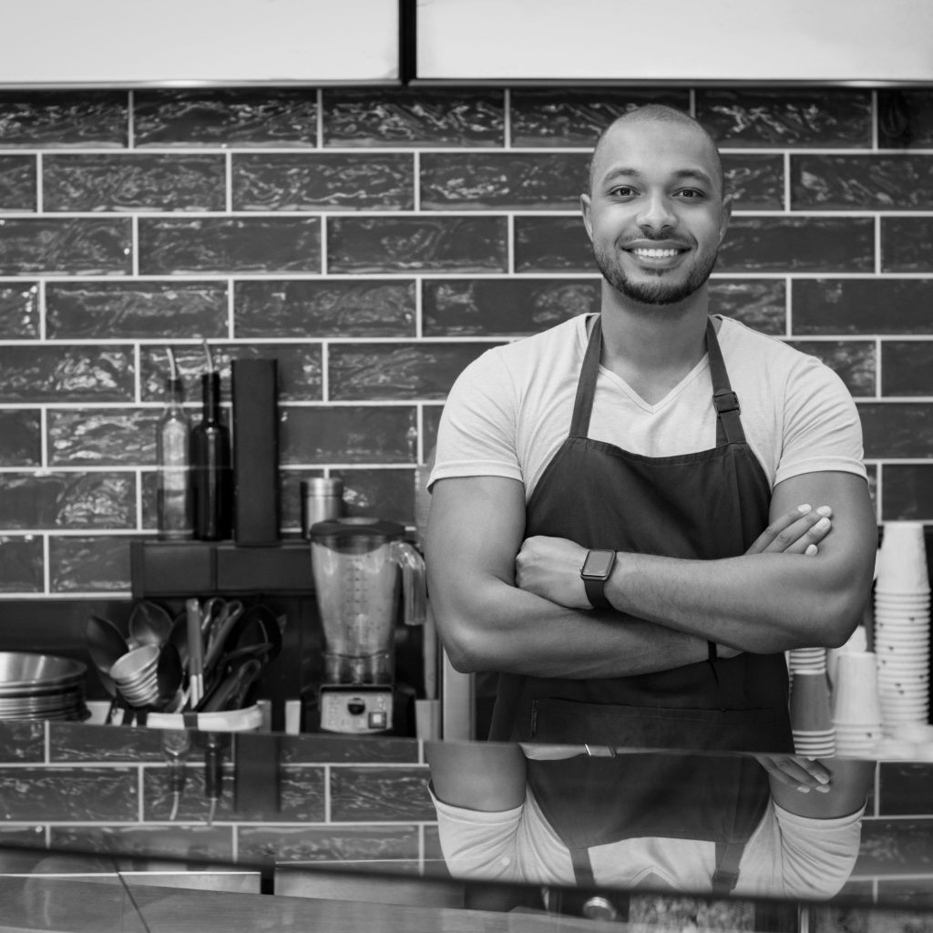 Smiling man in an apron stands behind a counter in a kitchen with a brick wall behind him.