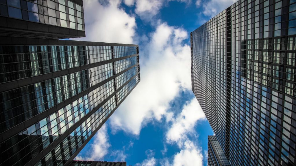 Two modern glass skyscrapers rise toward a blue sky with white clouds, viewed from ground level.