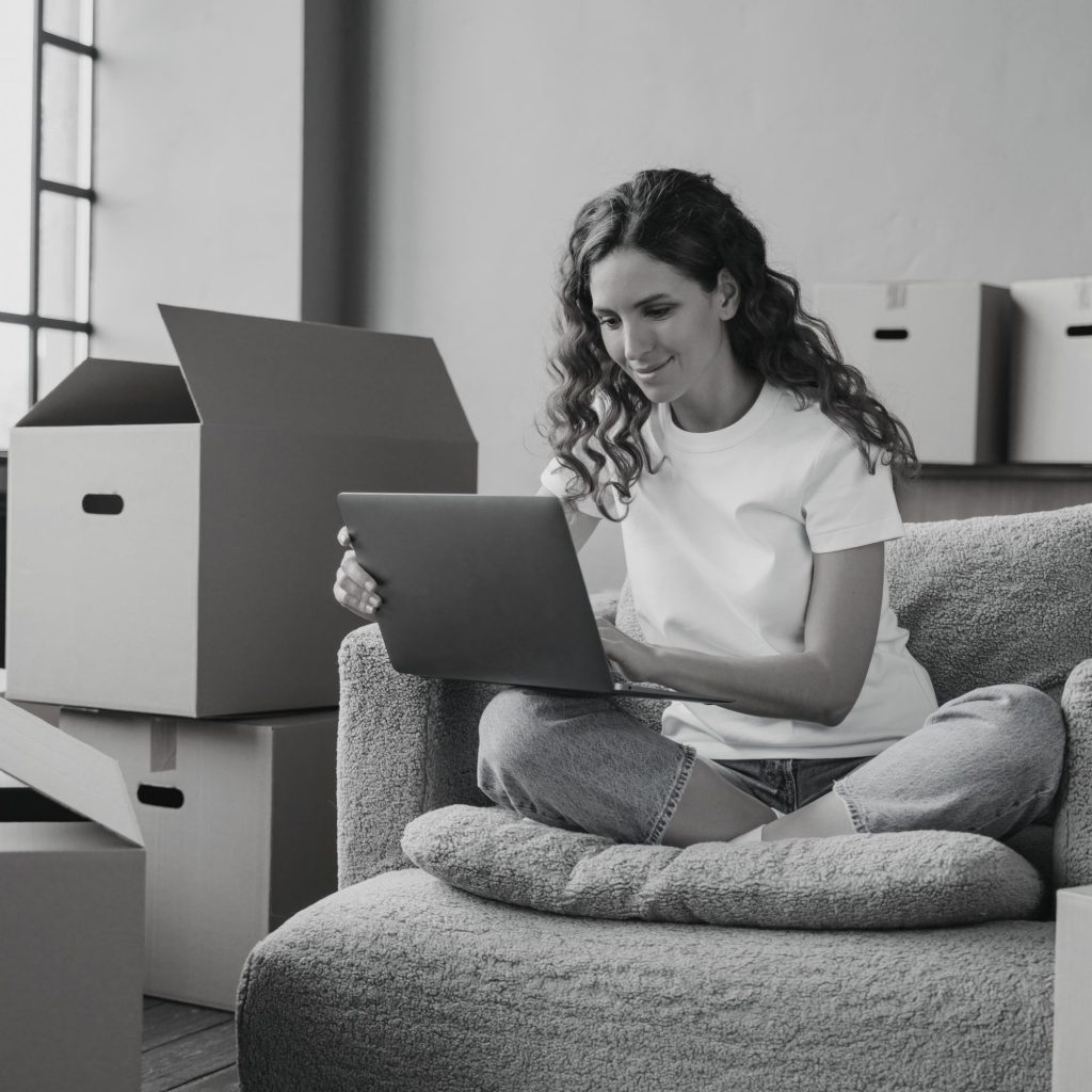 Woman sits on a fluffy sofa, using a tablet, with moving boxes around in a home.