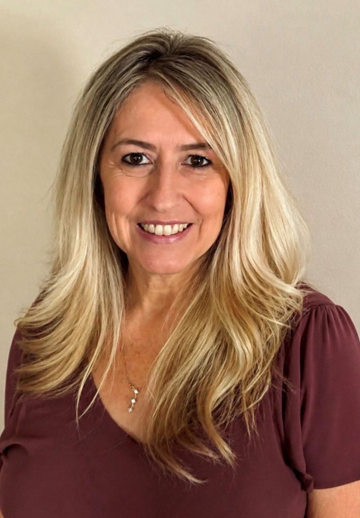 Smiling woman with long blonde hair in a maroon top, standing against a beige wall.